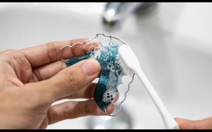 A close-up of a toothbrush cleaning a removable retainer under running water.